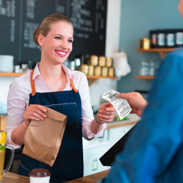 Sur la photo on voit une jeune femme avec un tablier derrière un comptoir. Elle tient un sac en papier dans sa main droite et elle sourit. À droite on voit une personne qui tient un ticket de caisse à la main. En arrière plan on voit des étagères avec des boites et un grand tableau noir avec de l'écriture blanc en craie.